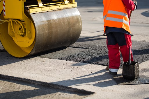 A worker in an orange vest and green pants shoveling asphalt.